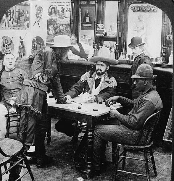 A man in a fringed leather jacket and cowboy hat holds a revolver at his waist as he argues with a seated fellow gambler in a staged photograph depicting a scene of drunken conflict during the Klondike Gold Rush, Alaska, early 1900s. (Photo by R.Y. Young/Hulton Archive/Getty IMages)